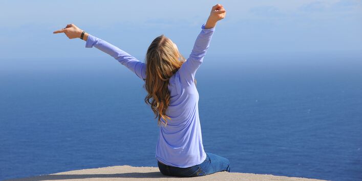 eine Frau sitzt am Meer und atmet tief durch, bis zum Horizont ist nur blaues Wasser zu sehen