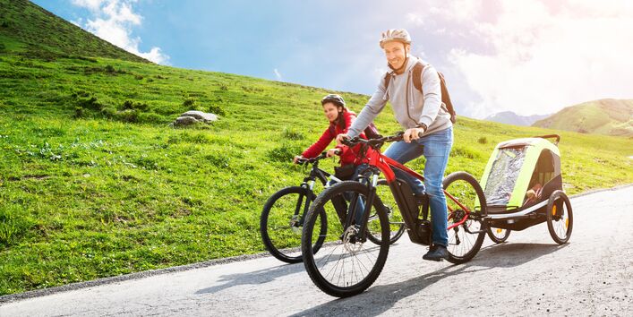 Mann auf einem E-Bike mit Kinder-Fahrradanhänger auf einer Straße in bergigem Gelände bei Sonnenschein