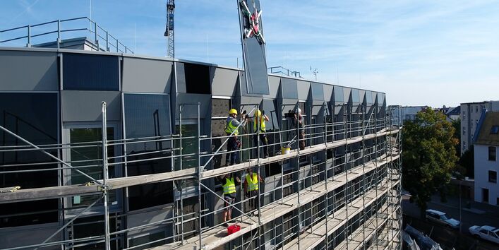eingerüstetes Gebäude mit Fassade aus Photovoltaikmodulen, Bauarbeiter auf dem Gerüst nehmen ein Modul in Empfang, welches an einem Kran hängt
