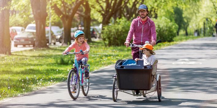 breiter und begrünter Radweg in der Stadt, ein Vater transportiert seine kleine Tochter in einem Lastenrad, eine größere Tochter fährt mit eigenem Rad nebenher