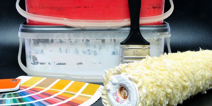 verschiedenfarbige Wandfarben in niedrigen Eimern, davor ein Fächer mit Farbmuster-Bögen, ein Pinsel und eine Rolle zum Wändestreichen