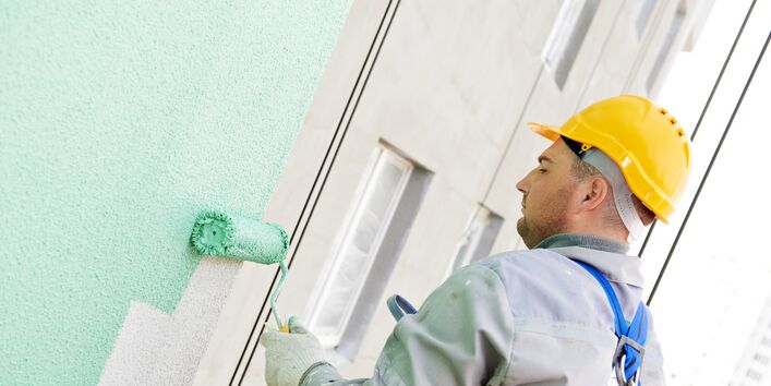 Man in overalls and yellow hard hat applying a mint green coat to a building façade