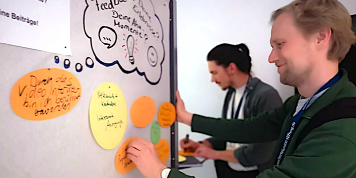 zwei Männer bringen Zettel an einer Stellwand zum Thema "Dein Feedback" an