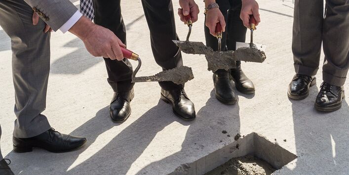drei Männer und eine Frau in festlicher Kleidung betonieren mit Maurerkellen eine quadratische Aussparung in einer Bodenplatte zu und lächeln in die Kamera