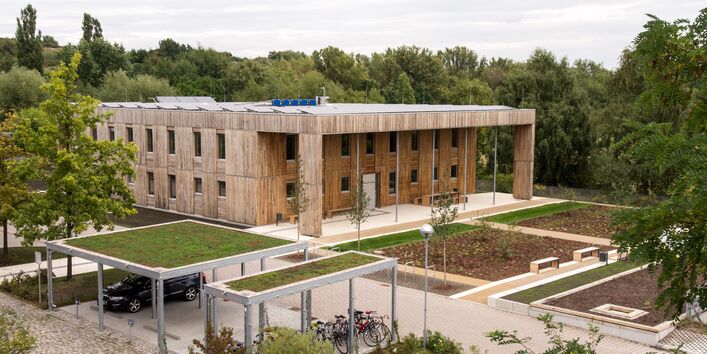 quadratischer, zweistöckiger Flachbau mit Holzfassade und Fahrradständerüberdachungen mit Dachbegrünung inmitten frisch angelegter und bepflanzter Außenanlagen