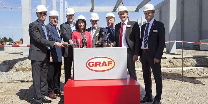 Sieben Männer und eine Frau haben sich anlässlich einer Grundsteinlegung mit Blauhelmen und Hämmern vor einem symbolischen Grundstein mit dem Firmenlogo "Graf" für ein Gruppenfoto aufgestellt 