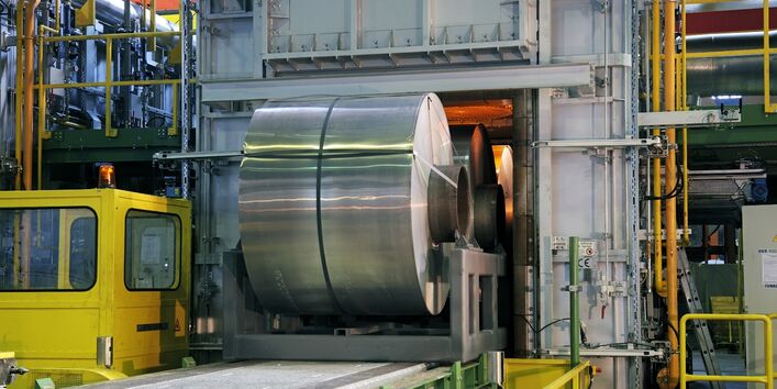 in einer Fabrikhalle wird ein große Rolle aufgerolltes Aluminiumband auf einem Förderband in eine Kammer gefahren, in der es glüht