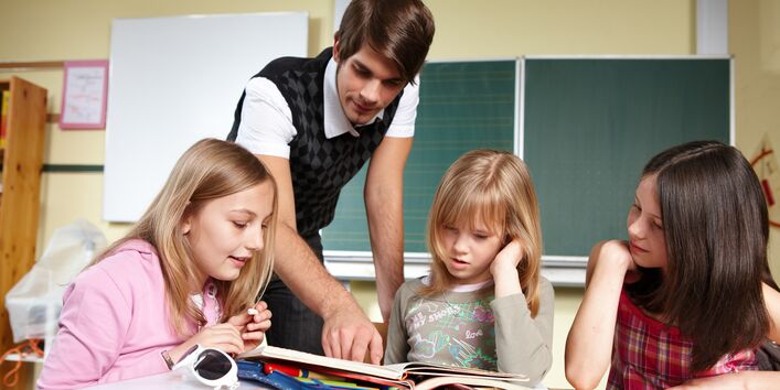 ein junger Leerer und drei Schulkinder sind an einem Tisch in einem Klassenzimmer über Hefte gebeugt