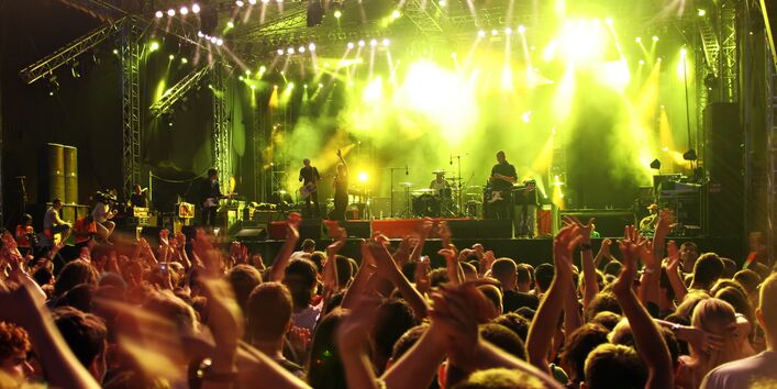 Menschen stehen dicht gedrängt und über dem Kopf in die Hände klatschend vor einer Konzertbühne, auf der eine Band im Scheinwerferlicht spielt