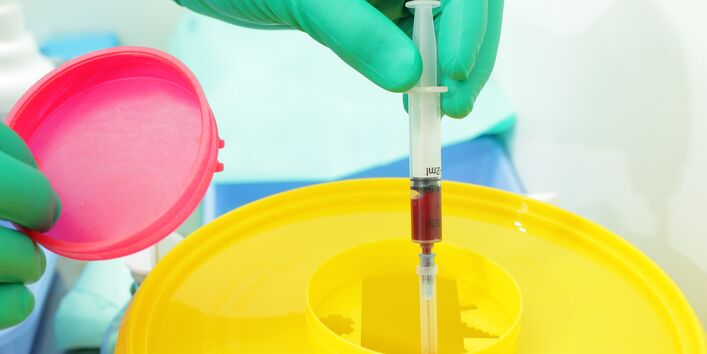 Hand mit medizinischem Handschuh tut eine benutzte Spritze in einen Behälter mit Deckel