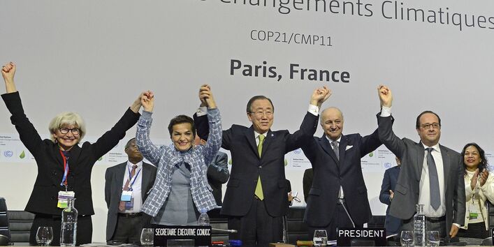 Women and men in suits hold hands and cheer, with the following text on the wall in the background: Nations Unies Conférence sur les Changements Climatiques COP21/CMP11 Paris, France