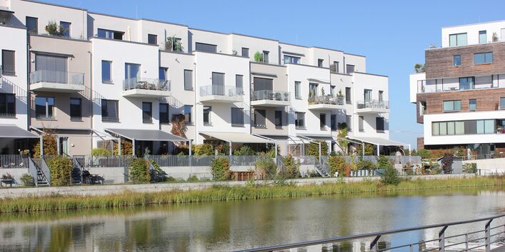an einem Kanal in der Stadt stehen weiße, vierstöckige Häuser im Bauhausstil mit Dachterrassen, Balkonen und kleinen Gärten mit Sonnenschutz