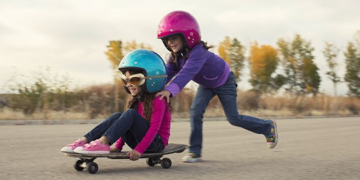 zwei kleine Mädchen mit Sturzhelmen schieben sich gegenseitig auf einem Skateboard auf einem gepflasterten Platz