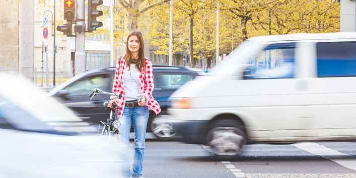 eine junge Frau wartet, ein Fahrrad schiebend, auf einer Mittelinsel an einer roten Ampel, vor und hinter ihr fahren Autos vorbei