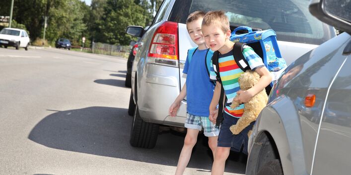 two children with satchels look between parked cars to see if they can cross the road safely