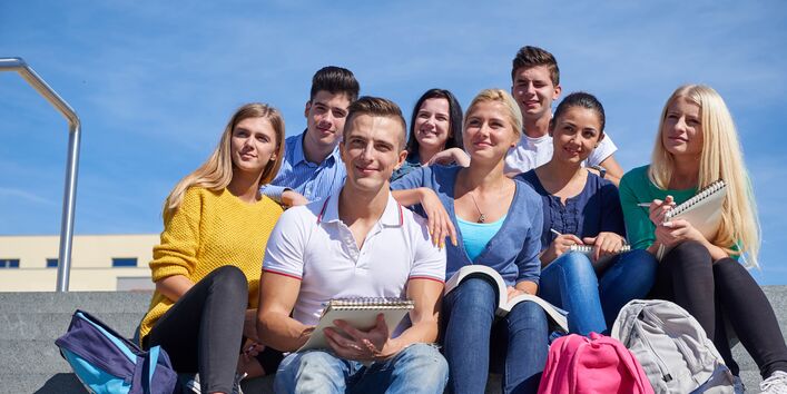 eine Gruppe Studentinnen und Studenten sitzt mit Rucksäcken und Schreibblöcken auf einer Steintreppe