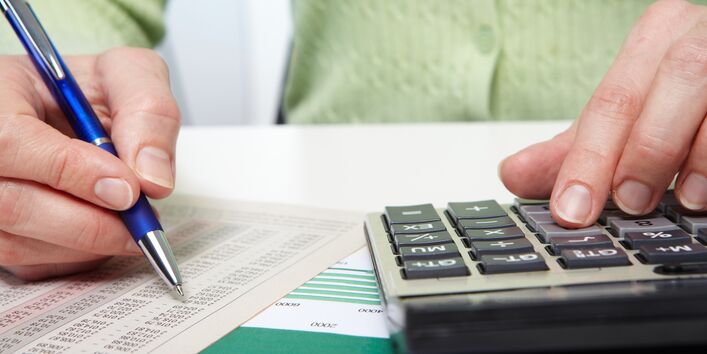 Frau sitzt an einem Schreibtisch und rechnet mit einem Taschenrechner, vor ihr liegt eine Liste mit vielen Zahlen