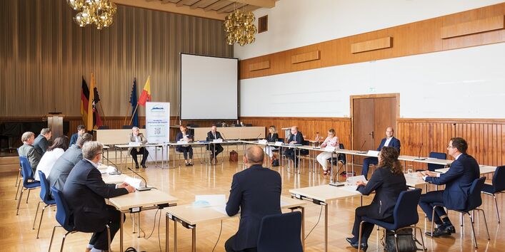 in einem Saal sitzen Männer und Frauen an Tischen im Kreis, hinten ein Aufsteller und Fahnen