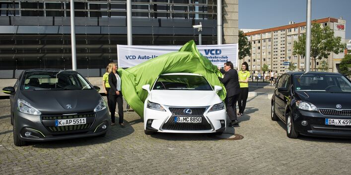 drei Autos stehen auf einem gepflasterten Platz, eine Frau und ein Mann ziehen ein grünes Tuch vom mittleren Auto, um es zu enthüllen. Im Hintergrund steht ein Banner des Verkehrsclubs Deutschland