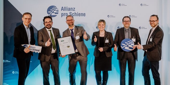 fünf Männer im Anzug und eine Frau im Kostüm stehen lächelnd mit einer Urkunde für ein Gruppenfoto postiert vor einer Stellwand mit dem Logo "Allianz pro Schiene"