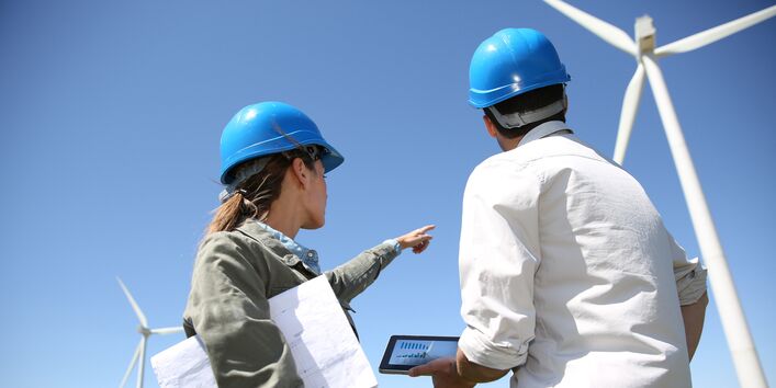 eine Frau und ein Mann mit Bauhelmen, Planunterlagen und einem Tablet schauen sich einen Windpark an