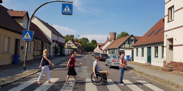 Straße mit niedrigen Fachwerkhäusern und einem Zebrastreifen, der von einer Frau mit Blindenstock, einem Rollstuhlfahrer und zwei weiteren Fußgängerinnen überquert wird.