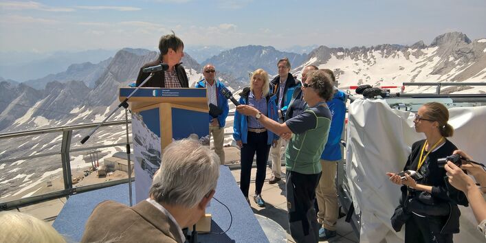 Barbara Hendricks steht auf einer Aussichtsplattform in den Bergen an einem Rednerpult mit Mikrofon, darum versammelt Reporter und andere Personen. Auf den umliegenden Gipfeln liegt Schnee.