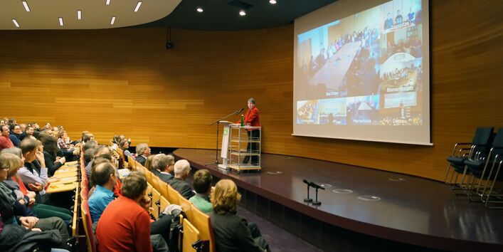 Barbara Hendricks hält im Hörsaal des UBA eine Rede, im Hintergrund sieht man auf einer Leinwand die Videoübertragung an andere Standorte. Im Vordergrund das Publikum in Dessau.
