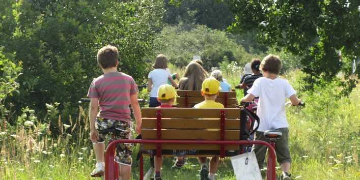 Eine Gruppe von Kindern sitzt auf einer Draisine auf Schienen und treibt sie mit Fahrradpedalen an.