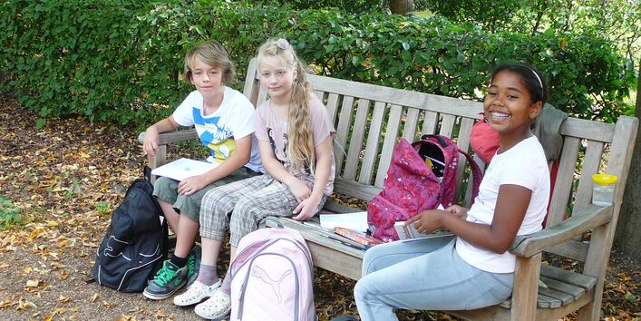 Drei Kinder sitzen auf einer Bank im Freien und malen, lachen