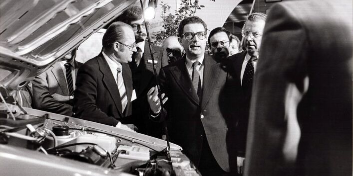 mehrere Männer stehen in einer Messehalle um ein Auto mit geöffneter Motorhaube herum.