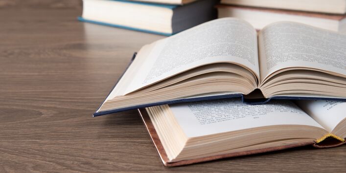 Zwei aufgeschlagene Bücher im Vordergrund, im Hintergrund zwei Bücherstapel