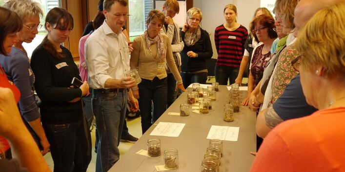 Menschen gruppieren sich um einen Tisch, auf dem Gläser mit verschiedenen Materialien stehen und die erläutert werden