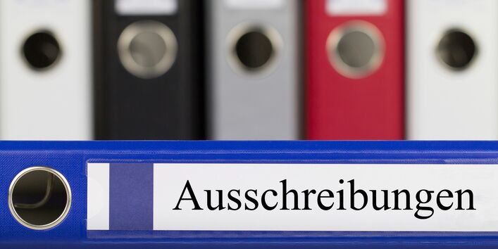 Mehrere Aktenordner im Hindergrund. Im Vordergrund ein blauer Aktenordner mit der Aufschrift Ausschreibungen.