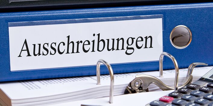 auf einem Schreibtisch liegen ein Ordner mit der Aufschrift "Ausschreibungen", ein Taschenrechner und ein Stift