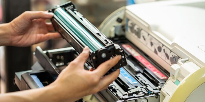 Jemand hält eine Tonerkartusche vor einem Druckgerät in der Hand