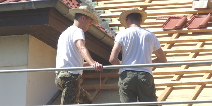 Zwei Männer mit Strohhüten auf Baugerüst beim Decken eines Daches.