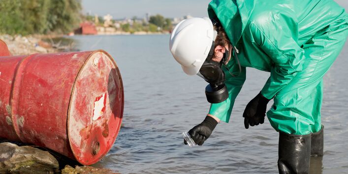 Ein rotes Fass liegt in einem Fluss und wird von einer Person in grünem Schutzanzug untersucht.