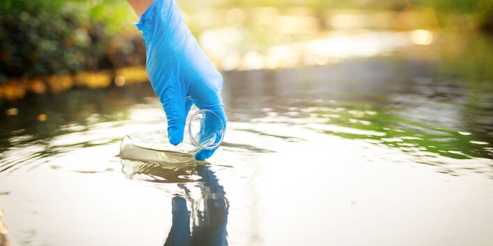Foto Hand mit Plastikhandschuh nimmt Wasserprobe in Glasgefäß aus einem Gewässer.
