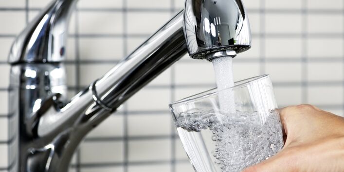 Ein Wasserhahn läuft und eine Hand mit einem Glas ist zu sehen.