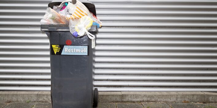 Eine überquellende Abfalltonne vor einem silberfarbenen Tor.