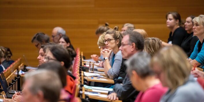 Sie sehen ein Bild welches auf der 1. Nationale Konferenz 2018: „Klimaanpassungsdienste – Klimavorsorge in der Praxis aufgenommen wurde. Sie sehen drei Reihen (von der Seite fotographiert) eines voll besetzten Hörsaals. Alle Menschen schauen sehr konzentriert und bis auf eine weibliche Person die gespannt nach vorne Blickt, sind alle Personen unscharf dargestellt.
