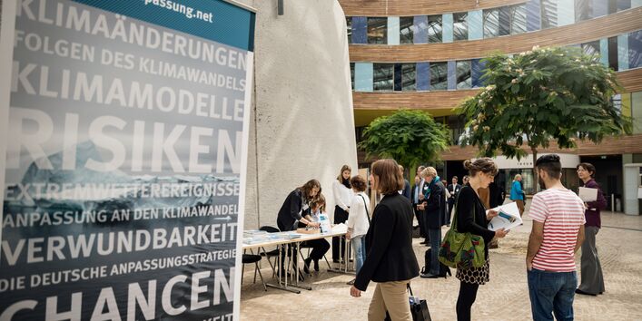 Sie sehen ein Bild vom Nationaler Dialog 2017 Gemeinsam sind wir klimaresilient – Wie Beteiligung in Städten und Regionen gelingt. Das Bild wurde im Umweltbundesamt in Dessau aufgenommen. Auf der rechten Seite sehen Sie ein Aufsteller zum Thema Klimarisiken und Anpassung. Auf der rechten Seite des Bildes sind Menschen auf dem Weg in den Hörsaal in Dessau.