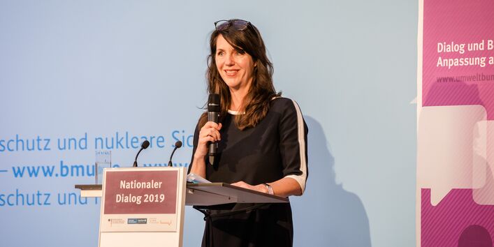 Dies ist ein Bild von dem Nationaler Dialog 2019: Klimawandelfolgen in Deutschland – Bereit zu handeln für bessere Vorsorge. Man siehr eine weibliche Perosn mit langen schwarzen Haaren eine Rede halten. Sie steht auf einer Bühne, an einem Pult und strahlt in die Kamera.