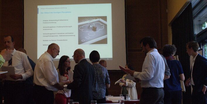 Ein Bild von dem Stakeholderdialog 2019: Katastrophenvorsorge bei Wasserengpässen kooperativ gestalten. Man sieht im Hintergund die PowerPoint Präsentation auf der das Planspiel erklärt ist, welches im Vodergrund abläuft. Personen stehen in kleineren Gruppen zusammen und diskutieren miteinander. 