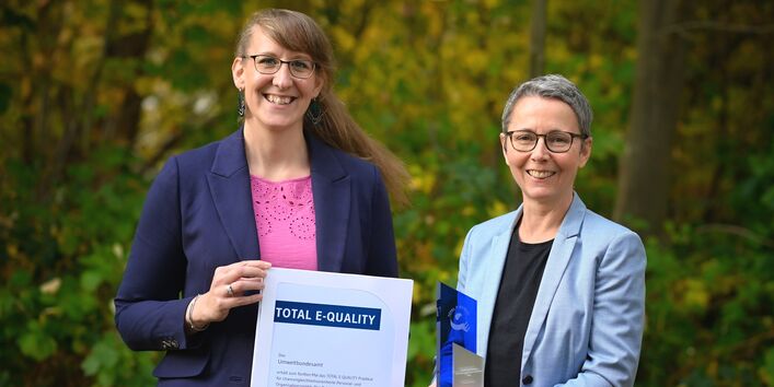 zwei freundlich lächelnde Frauen präsentieren das Zertifikat und die Trophäe 