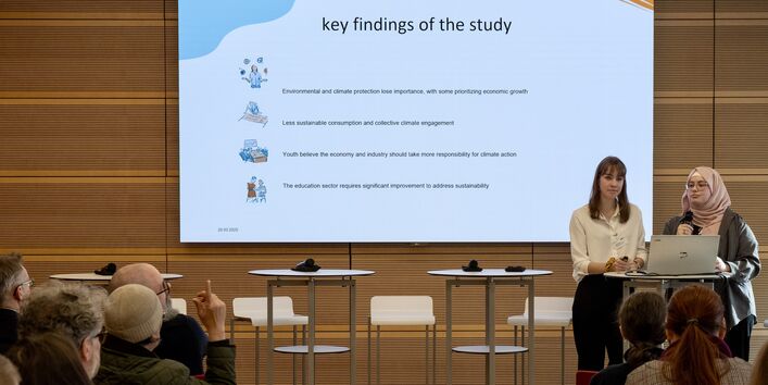 Präsentation auf dem Bildschirm auf der Bühne. Zwei Damen sprechen dazu an einem Stehtisch.