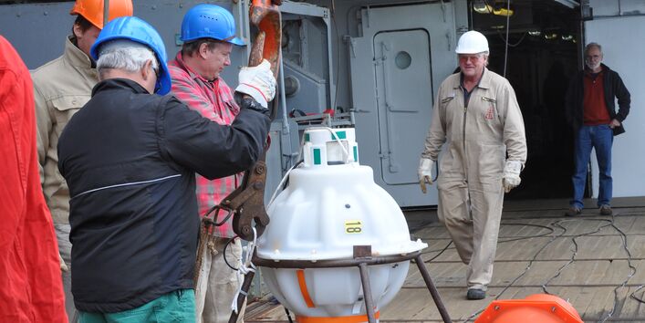 An Bord der Polarstern bereiten 5 Männer die PIES-Sonde auf ihren Einsatz vor. Die Sonde ist rundlich und hängt ein einem Kran, der sie zum Meer hinablässt. 