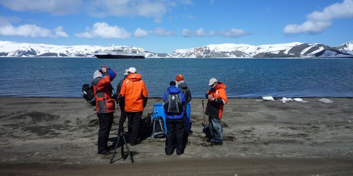 Reisende stehen im Vordergrund auf Land. Im Hintergrund sieht man antarktisches Meer und ein großes Schiff vor beschneiten Hügeln. 