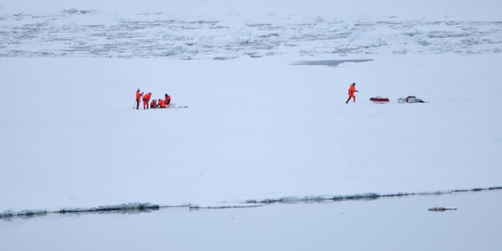 Auf einer weiten Eisebene steht eine Gruppe von Forschern und roten Kälteschutzanzügen. Sie entnehmen an einer Stelle eine Eisprobe. Weiter rechts läuft eine weitere Person zur Ausrüstung. 